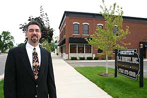 Man in a suit standing on a sidewalk with a building and a sign in the background.