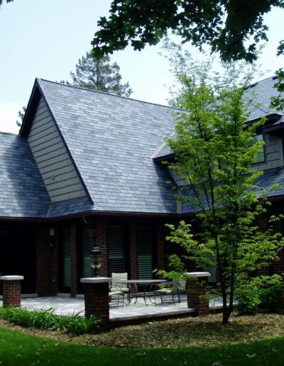 A brick house with a gray shingle roof surrounded by lush greenery.