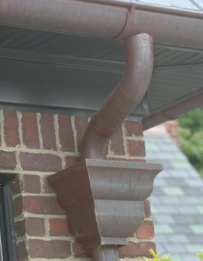 A close-up view of a brown gutter downspout attached to a red brick house with green foliage in the background.