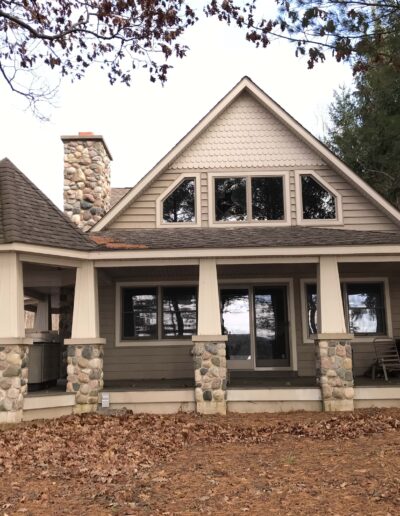 A single-story house with a stone foundation, a covered porch, and an a-frame design amidst a backdrop of trees.