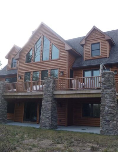 A large two-story wooden house with stone foundation accents and a balcony, surrounded by trees.